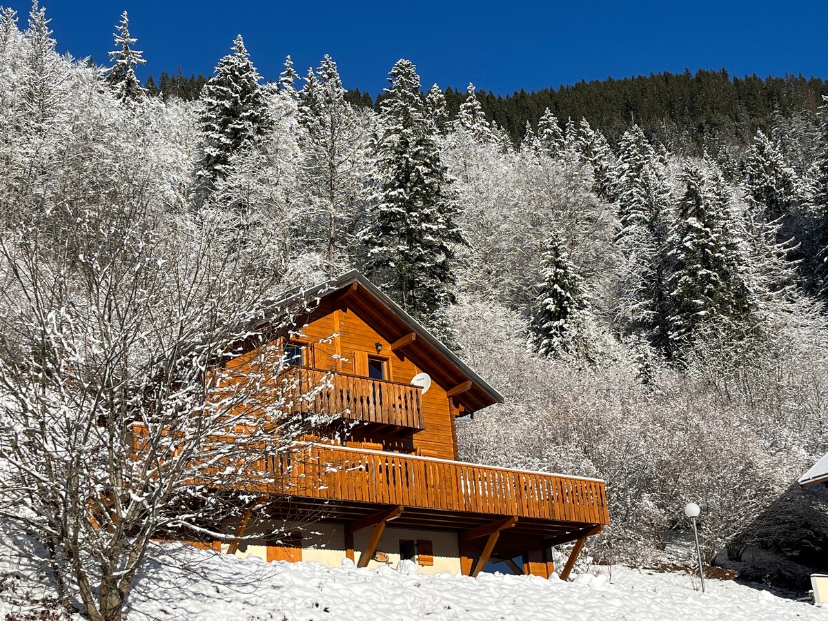 A wooden chalet stands amidst a snowy landscape, surrounded by evergreen trees. A balcony extends from the upper floor, offering views of the serene setting. The structure's warm tones contrast with the crisp white snow covering the ground and nearby flora.