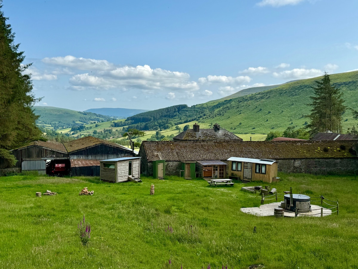 A serene landscape is captured, featuring a lush green meadow surrounded by rustic structures. The tranquil setting is framed by rolling hills under a clear blue sky, with a wood-fired hot tub and seating area visible in the foreground, inviting relaxation in nature.