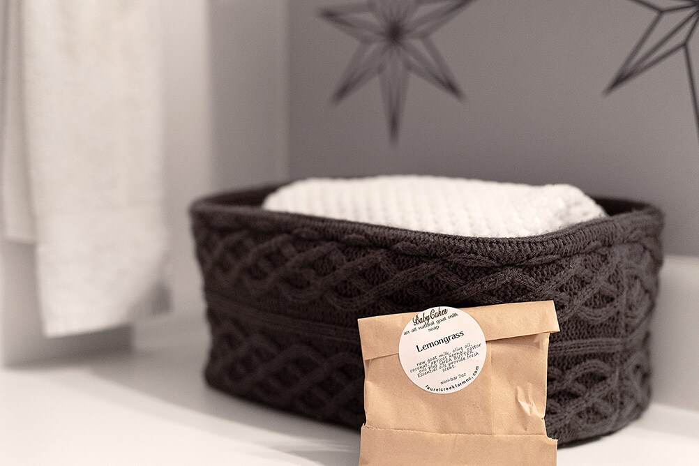 A textured black woven basket holds a white towel, resting beside a small package labeled 'Lemongrass.' The background features a subtle design with stars, contributing to a calming and organized atmosphere.
