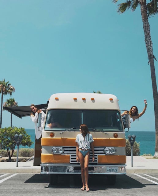 A vintage motorhome is prominently displayed in a coastal parking area, with palm trees framing the background. Bright sunlight complements the retro design, showcasing an inviting outdoor space. People can be seen interacting near the open windows of the vehicle, with the ocean visible in the distance.