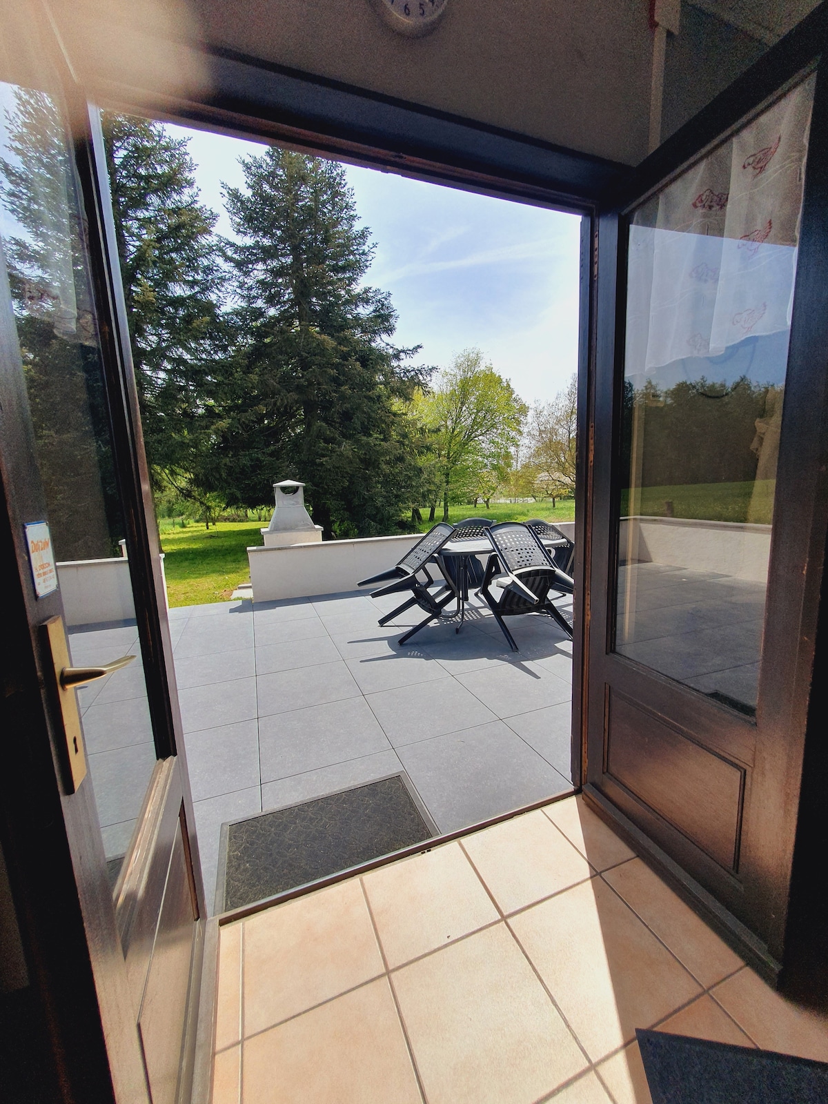 An open doorway leads to a spacious terrace featuring several black outdoor chairs arranged around a table. The terrace is paved and overlooks a large, green lawn with trees in the background, providing a serene outdoor environment.
