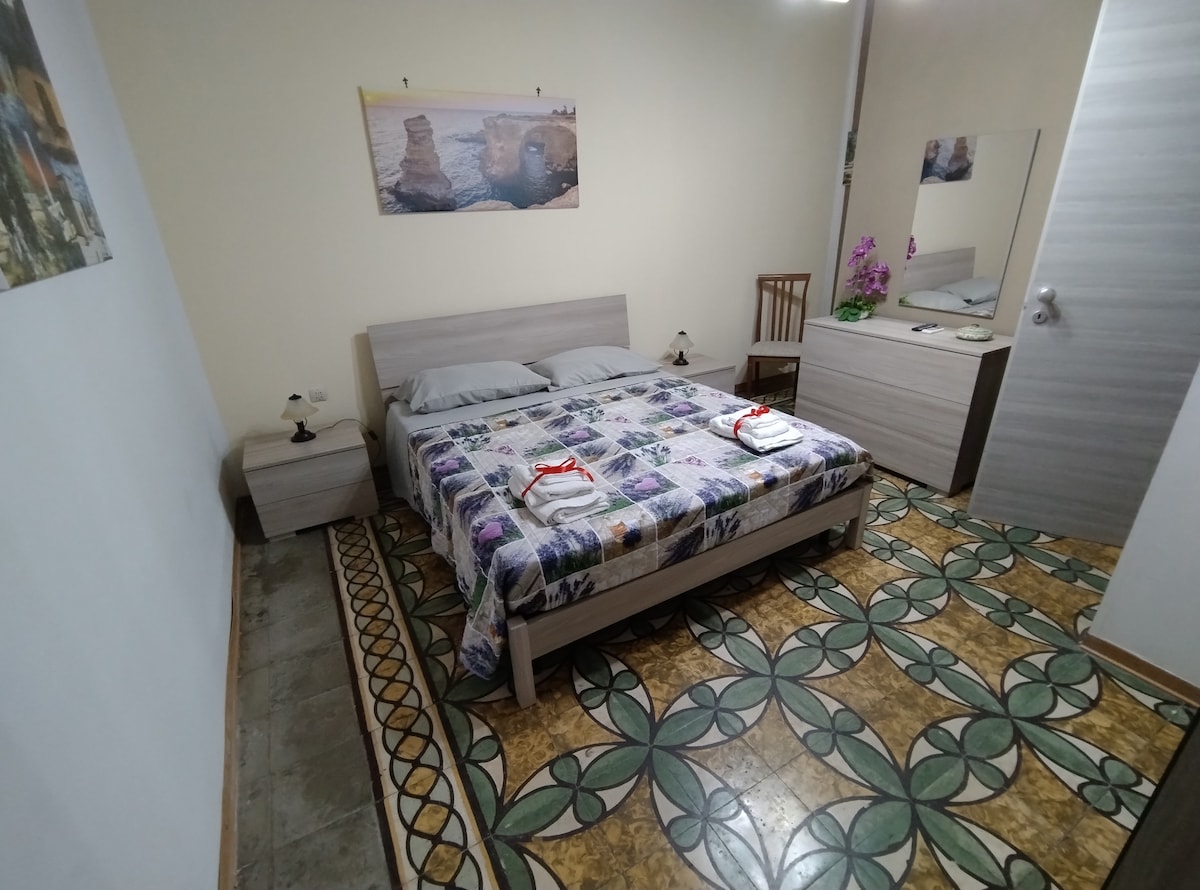 A light-filled bedroom showcases a spacious bed adorned with a floral-patterned blanket. Flanking the bed are two nightstands with lamps. A mirror and a small chair are positioned near the wall, while a piece of artwork hangs above the bed, adding a personal touch to the space.