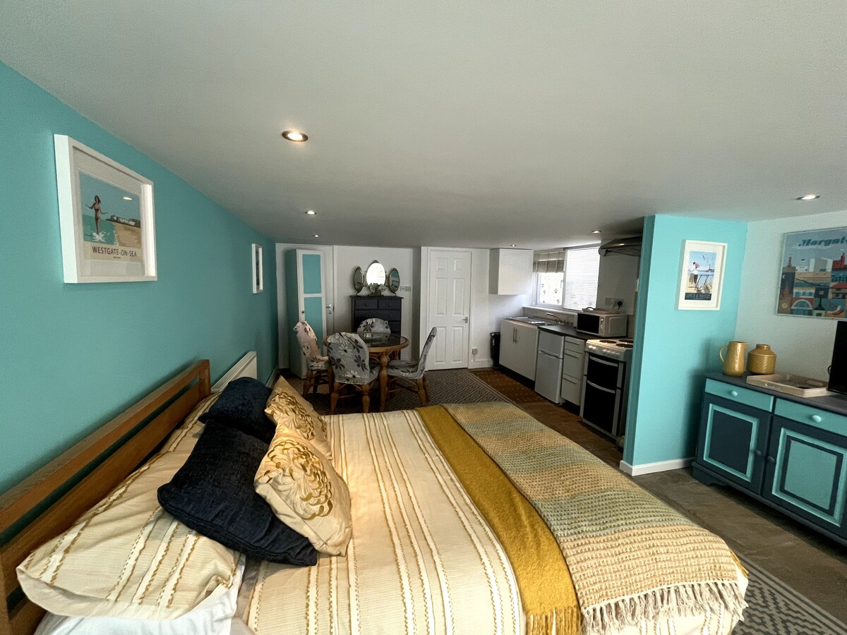 A spacious studio summerhouse features a comfortable sleeping area with a warm-coloured blanket draped over the bed. The room is accented by turquoise walls and contains a small dining table and chairs, along with a fully equipped kitchen visible in the background.