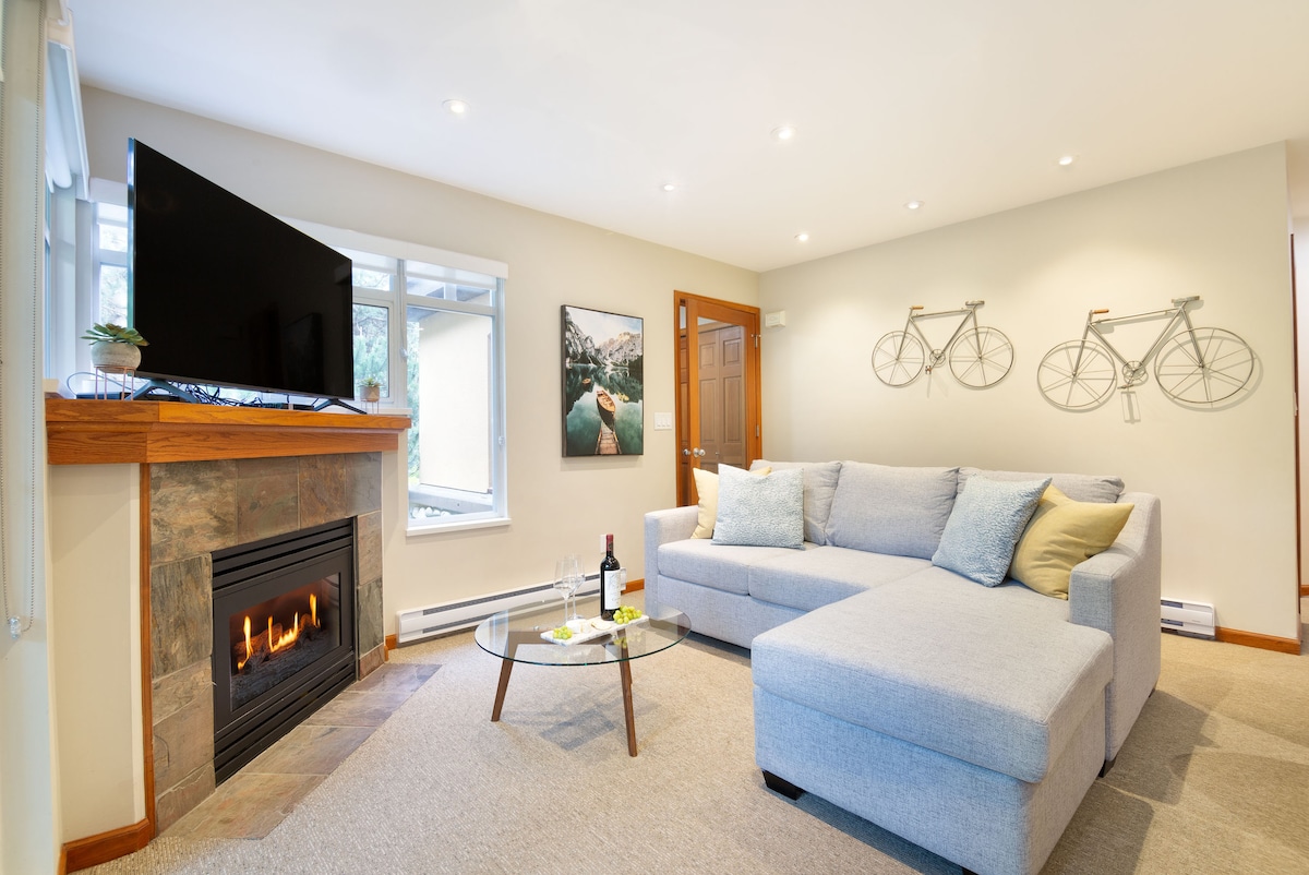 A cozy living area features a spacious sectional sofa faced towards a large television, which is mounted above a stone fireplace. Natural light filters through a nearby window, highlighting a wall adorned with bicycle art.