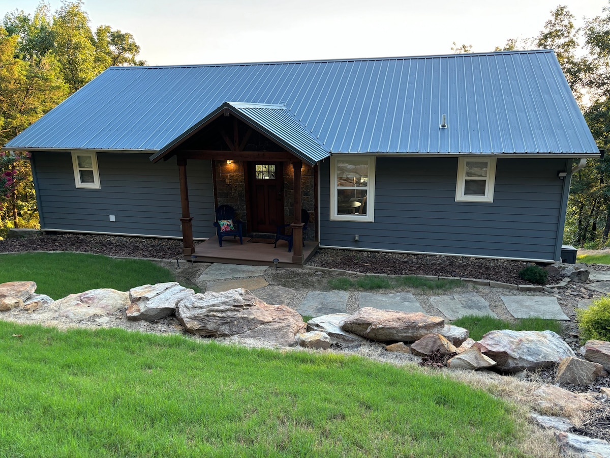 The exterior of the cozy mountain cottage is shown, featuring a welcoming entrance with a covered porch. The house is surrounded by green grass, colorful landscaping, and rock features, complemented by a metal roof that reflects the sunlight.