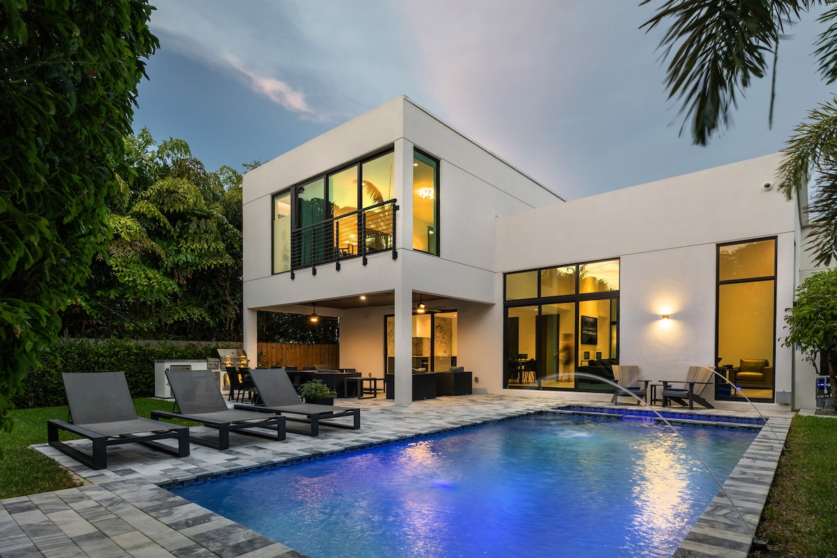 The exterior view of a modern home reveals a spacious backyard featuring a heated swimming pool with illuminated water. Surrounding lounge chairs provide relaxation areas. The house showcases large glass windows and an outdoor dining space, framed by lush greenery.