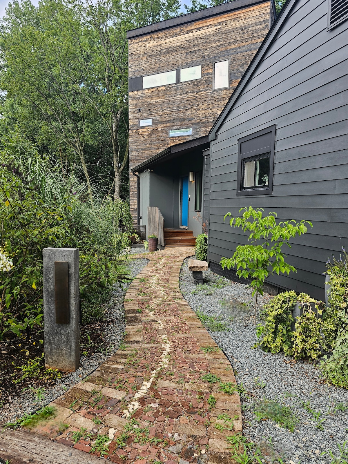A winding brick pathway guides visitors toward the entrance of the Retreat, bordered by lush greenery and gravel. The exterior of the building features a mix of dark wood and siding, with a bright blue door that adds a visually appealing contrast.