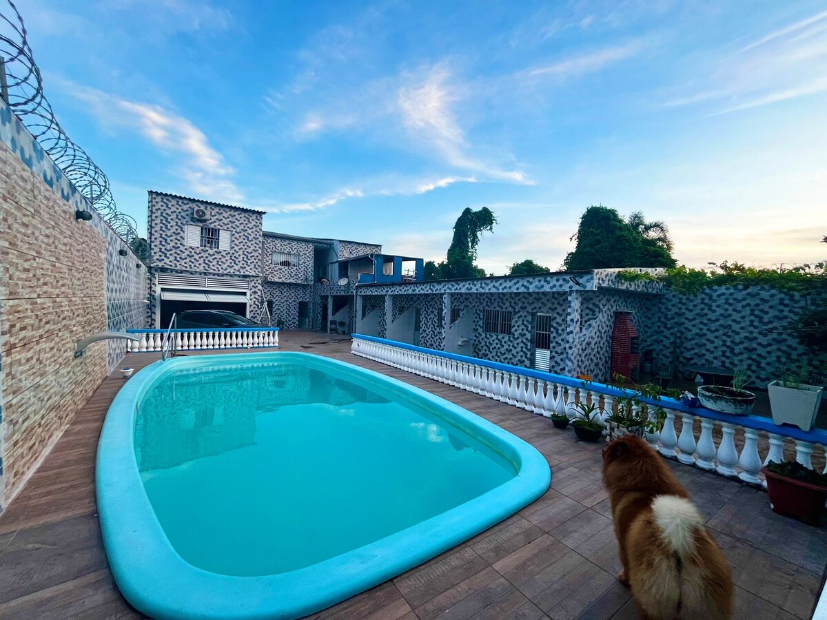 A shared swimming pool is positioned in the foreground, surrounded by a tiled deck. At the back, several buildings with a unique patterned facade are visible. Lush greenery is present along the periphery, complementing the outdoor space.