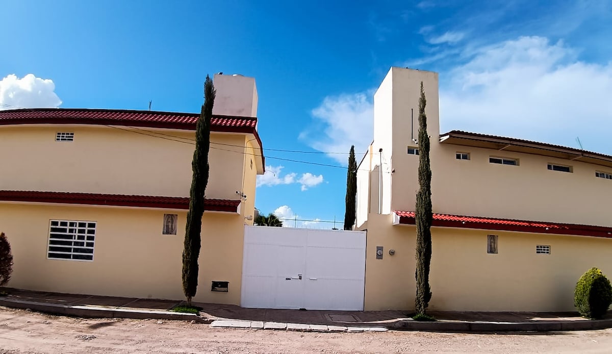 A modern apartment building features a light-colored exterior with a red-tiled roof. Tall cypress trees frame the entrance, which is marked by a large white gate. The sky is bright with a few clouds visible overhead.