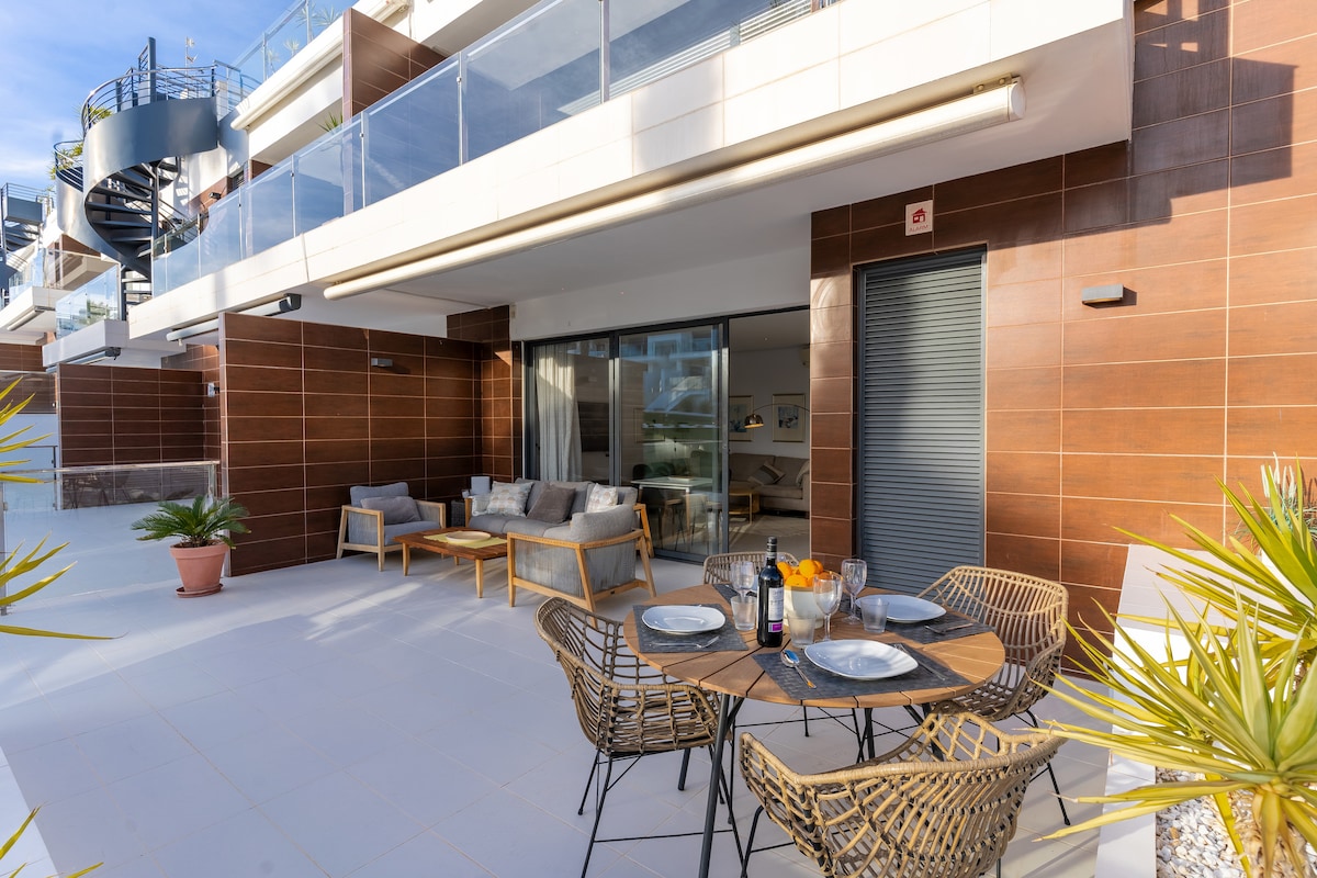 An outdoor terrace features a round dining table set with plates and glasses. Several wicker chairs surround the table. The space is complemented by a seating area with two sofas, and large sliding glass doors provide a view into the living area.
