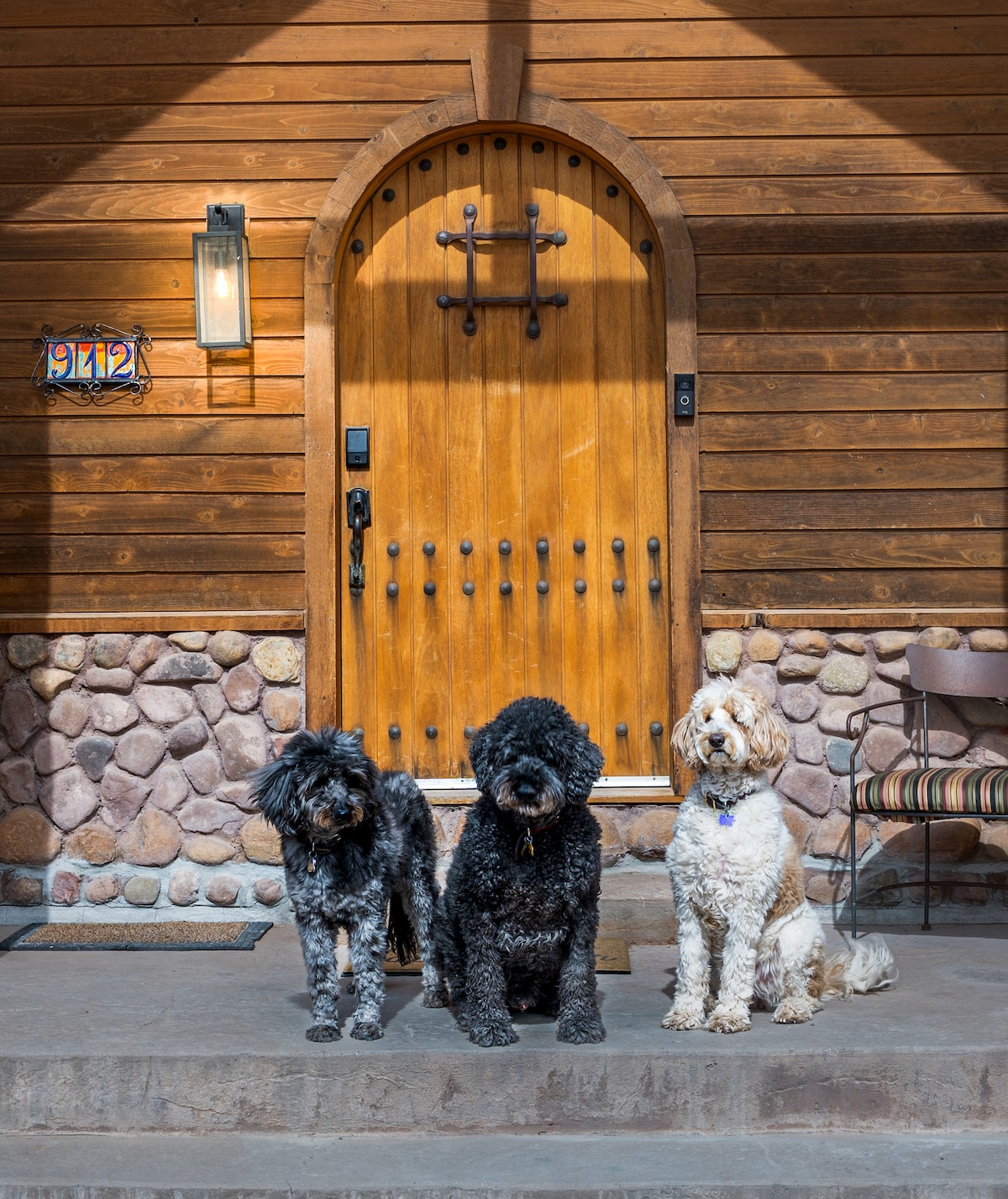 Three dogs are positioned on a stone-fronted porch in front of a wooden door. The door features an arched design with a wooden cross and a small window. Natural wood tones of the house complement the decorative elements.