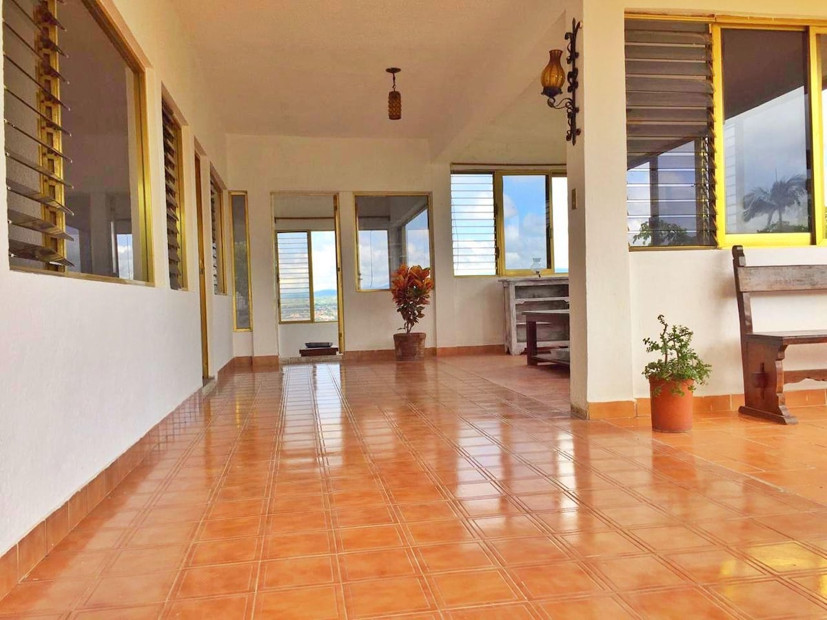 An open and airy corridor features polished tile flooring and large windows allowing natural light to fill the space. Potted plants add a touch of greenery, while comfortable seating is arranged in a corner. The views beyond the windows enhance the inviting ambiance.