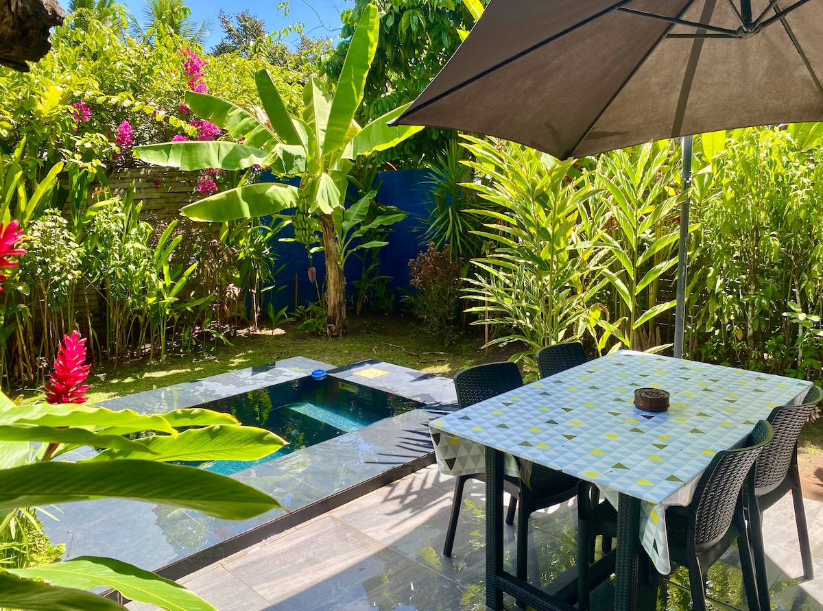 An outdoor dining area is set under a large umbrella, featuring a table surrounded by four black chairs. A small private pool is visible nearby, with lush tropical foliage and vibrant flowers enhancing the serene garden atmosphere.