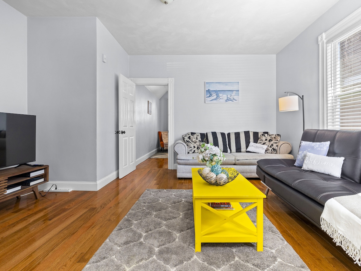 A bright living room features a mix of seating options, including a gray couch and a dark leather sofa. A vibrant yellow coffee table sits atop a soft area rug, complemented by decorative items. Large windows allow natural light to enhance the space.