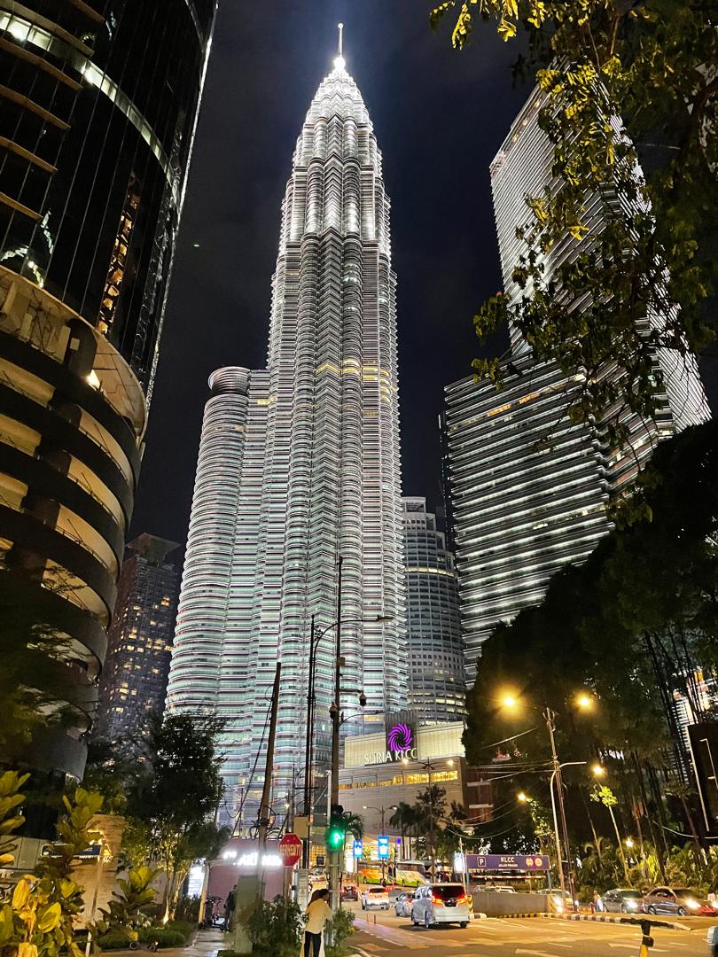 The Petronas Twin Towers are illuminated at night, showcasing their iconic modern architecture. The towers rise prominently above nearby structures, reflecting city lights. Surrounding palms and greenery enhance the urban landscape, while the street below is lined with vehicles and glowing street lamps.