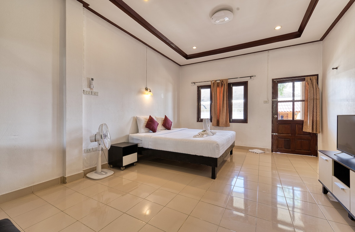 A spacious room featuring a king size bed with a single bed beside it. Natural light enters through two windows, illuminating the tiled floor. A wooden door leads to the entrance, and a fan is positioned in the corner for added comfort.