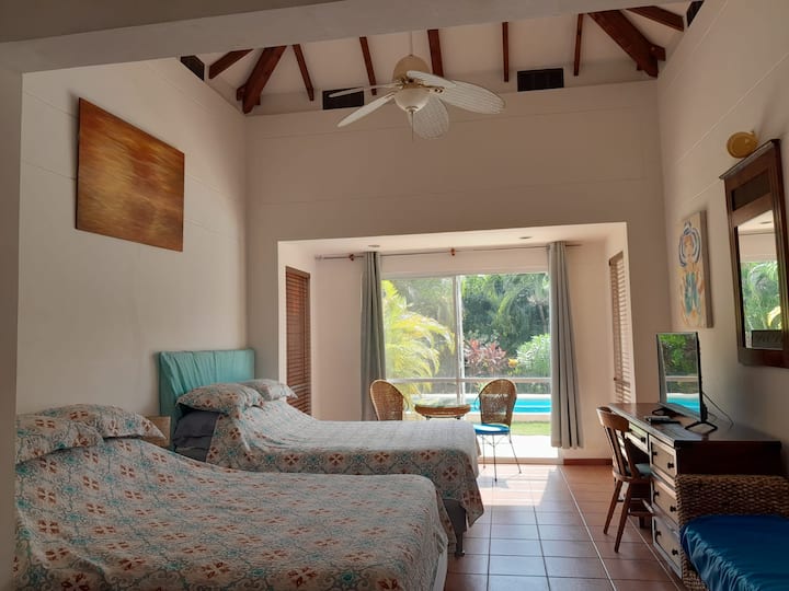 El dormitorio 2 ofrece dos camas tamaño queen bajo un techo alto con vigas de madera a la vista. Las grandes puertas correderas de cristal se abren a un patio privado con vistas a la piscina. Un escritorio de madera con un televisor de pantalla plana y un reproductor de DVD añade comodidad y entretenimiento.