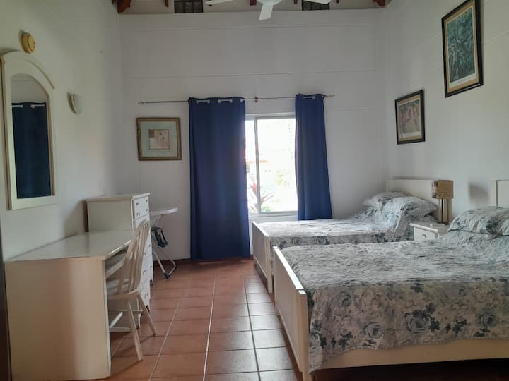 El dormitorio 3 cuenta con dos camas de tamaño completo, colocadas contra paredes blancas y baldosas de terracota. Una ventana con cortinas de color azul oscuro deja entrar la luz natural, mientras que un escritorio, un espejo y un ventilador de techo aportan calidez, limpieza y comodidad.
