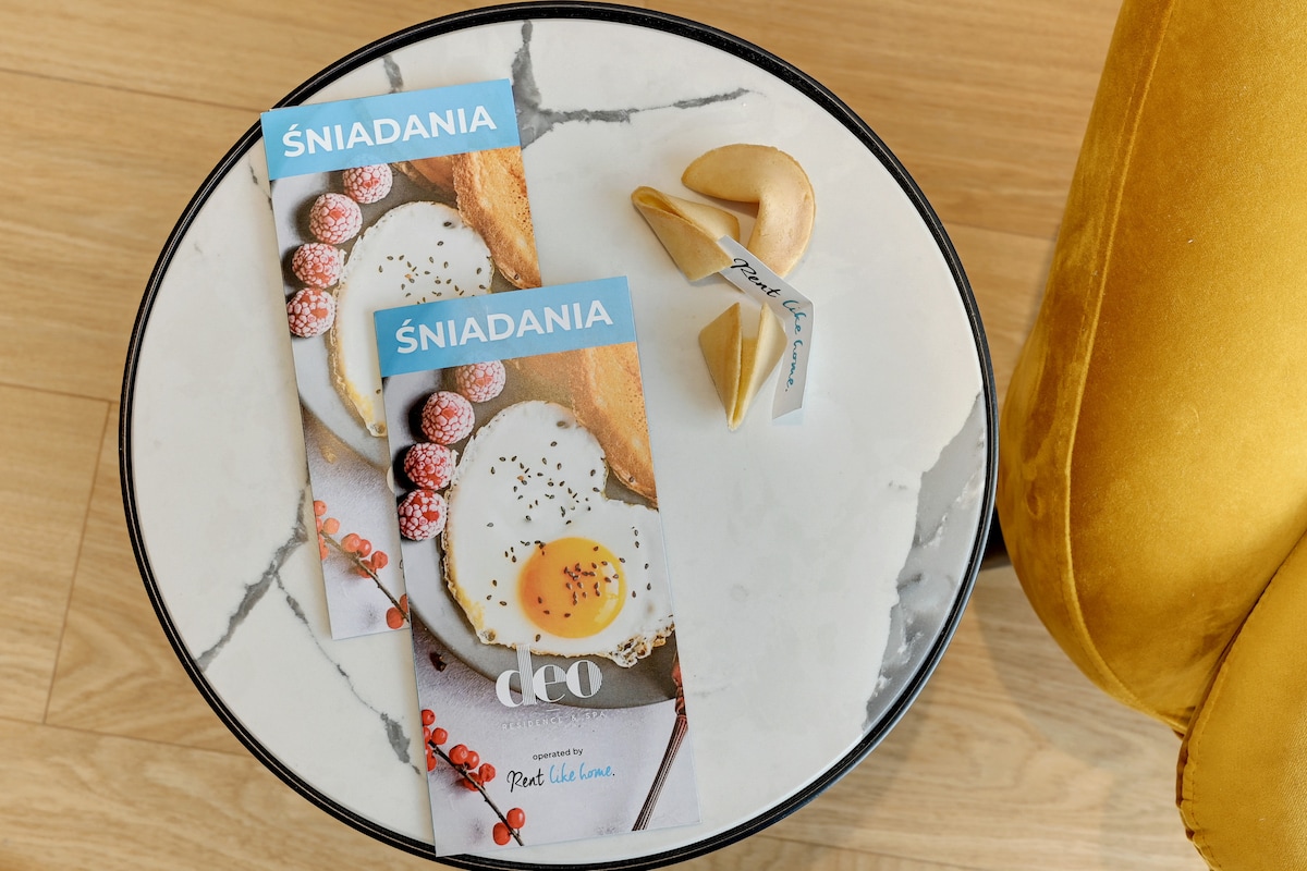 A marble-topped table displays two breakfast menus, featuring a soft-boiled egg and garnished with berries. Next to the menus, fortune cookies are placed, adding a playful touch to the setting. A vibrant yellow armchair is positioned nearby.