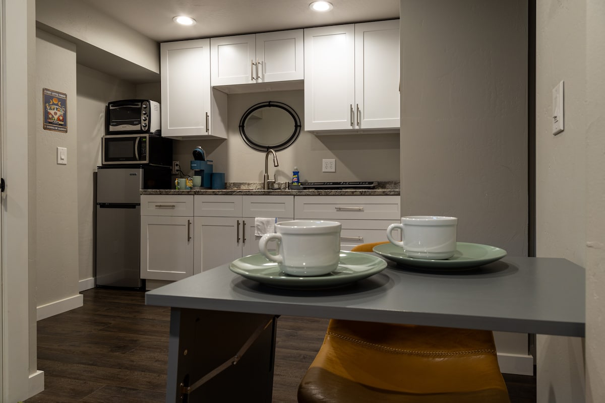 A small dining area is presented with a gray table and two chairs, each set with a mint green plate and white cup. The compact kitchen cabinetry and appliances are visible in the background, featuring white cabinets and dark countertops.