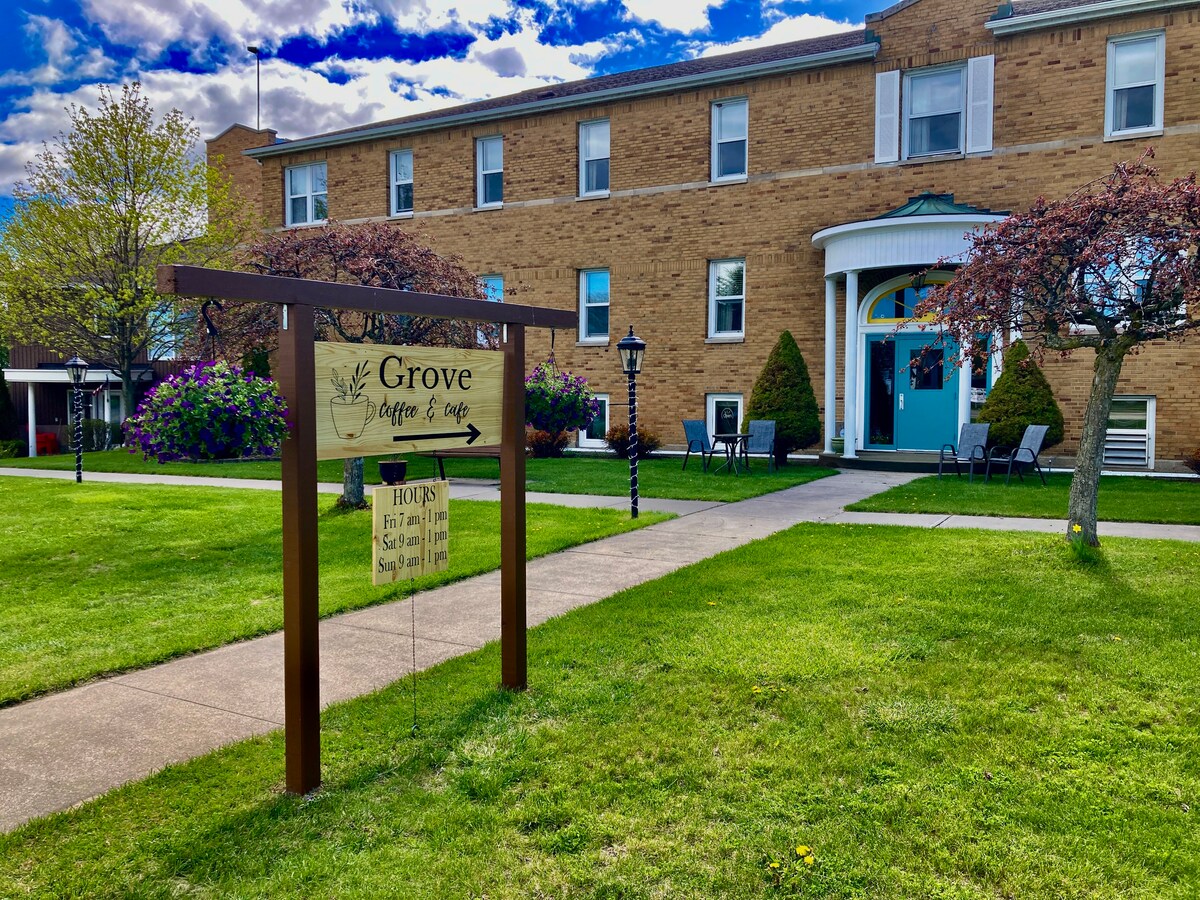 The entrance to the Grove Coffee & Cafe is seen, featuring a wooden sign in front of a charming brick building. Lush green grass surrounds the walkway, which leads to blue doors. Colorful flowering shrubs and trees line the pathway, contributing to the inviting atmosphere.