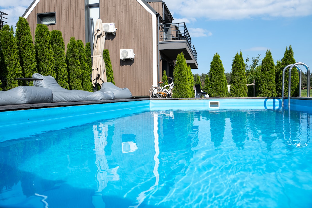 A seasonal swimming pool is featured, with clear blue water reflecting the sky. Surrounding greenery provides a sense of privacy. A shaded lounge area and sun loungers are visible, along with a bicycle parked in the background near the building's facade.