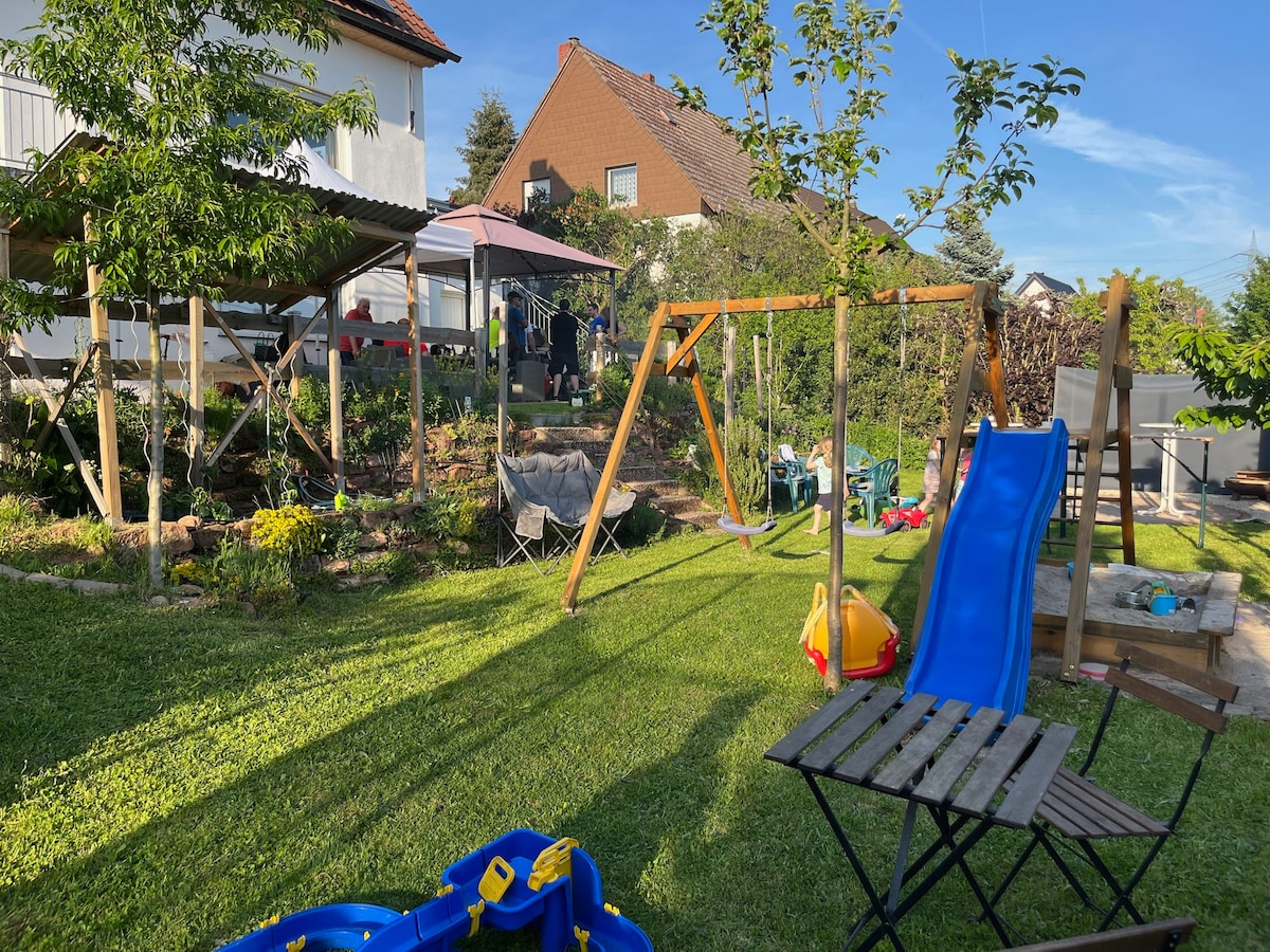 A spacious garden area features a playground setup, including a slide and swings. Lush green grass surrounds various outdoor seating options, with trees providing partial shade. In the background, a gazebo and additional recreational equipment can be seen, creating an inviting outdoor space.