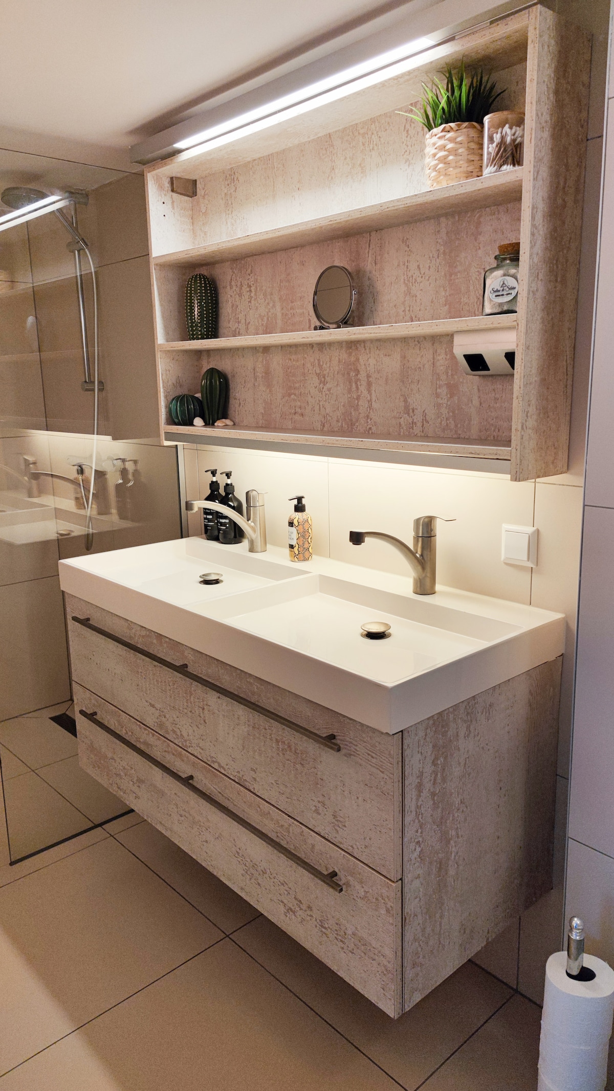 A modern bathroom features a double sink vanity with a smooth white countertop and wooden drawers below. Above the sink, a shelf displays various decorative items, including plants and toiletries. Soft lighting provides illumination within the space, enhancing visibility.