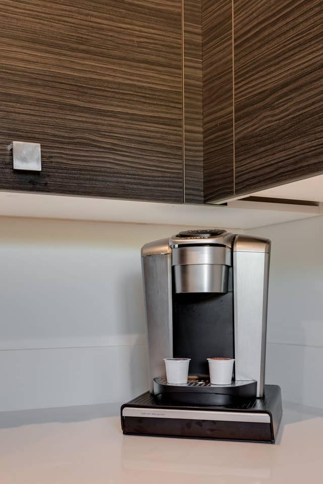 A modern coffee machine is positioned on a smooth countertop. Two small cups are placed beneath the machine's spout. Dark, textured cabinetry is visible above, complementing the sleek design of the kitchen area.