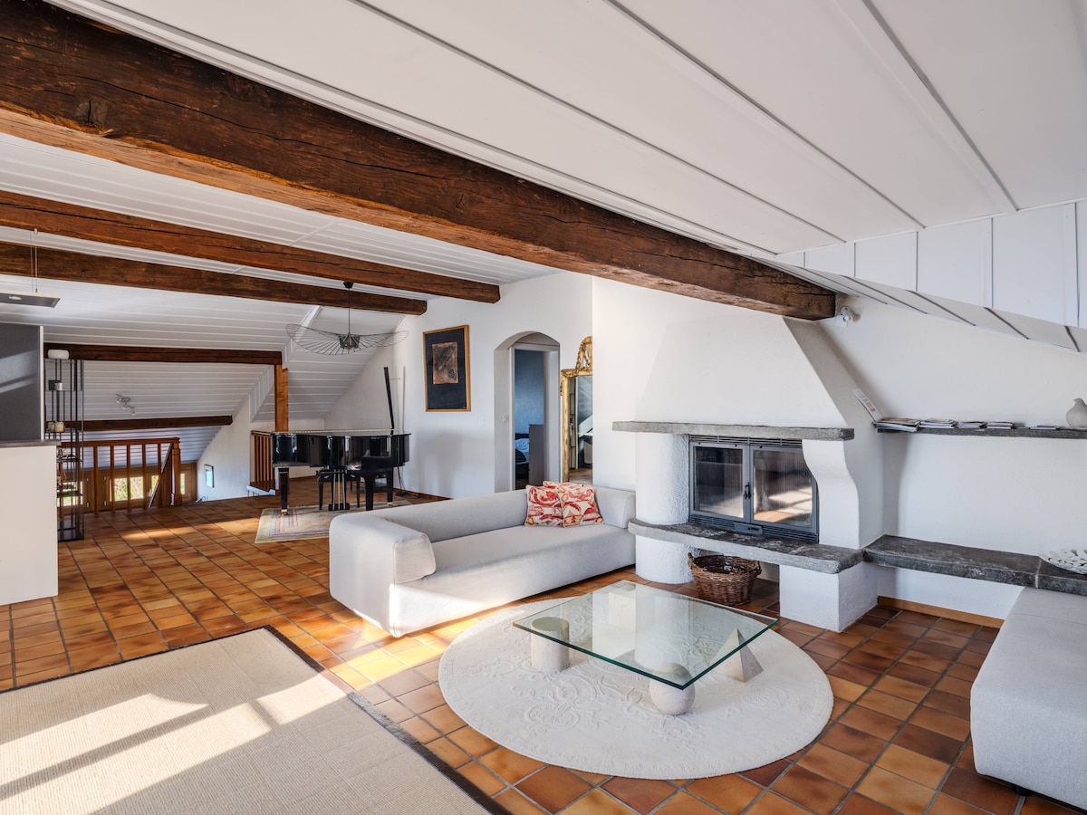 An airy living area features exposed wooden beams and a cozy fireplace. A sectional sofa is positioned around a glass coffee table, complemented by a light-colored area rug. Natural light enters through the windows, illuminating the terracotta tiled floor.