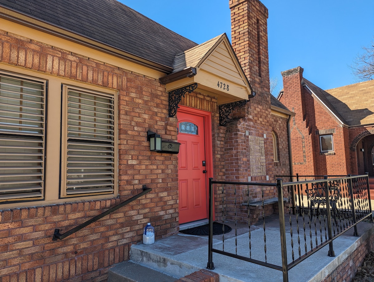 The exterior of a brick home is shown, featuring a bright orange front door with decorative accents. A short set of steps leads to the entrance, which is framed by a black wrought iron railing. The home displays a welcoming and well-maintained appearance.