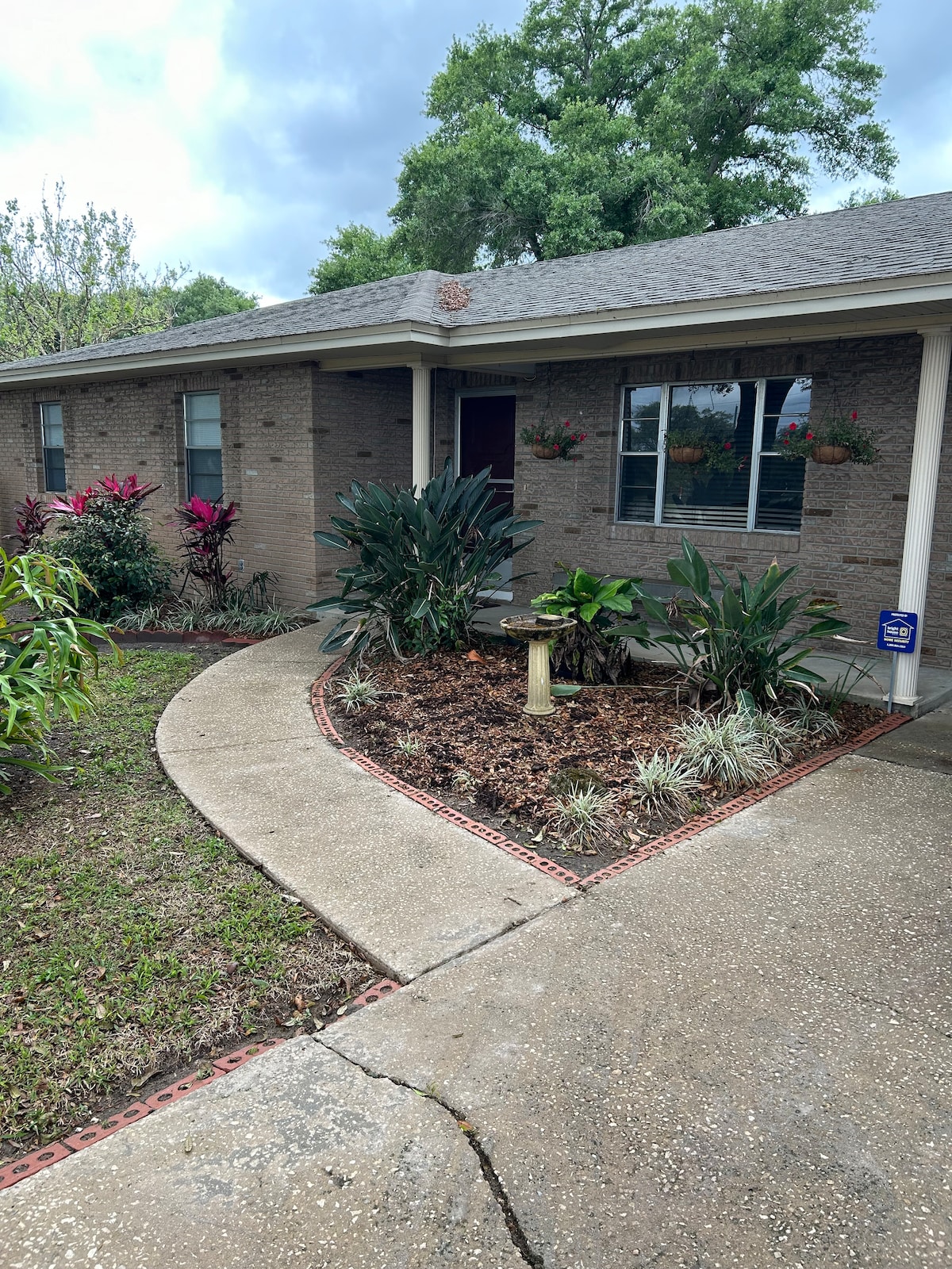 A landscaped entrance features a curved walkway leading to a brick facade. Lush green plants and colorful flowers border the path, enhancing the inviting atmosphere. A small birdbath is positioned among the foliage, complementing the serene garden setting.