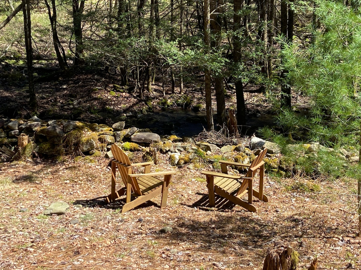 Two wooden Adirondack chairs are positioned outdoors, surrounded by a natural environment with sun-dappled light filtering through trees. A meandering stone wall covered with moss is visible in the background, alongside a gently flowing stream, creating a serene spot for relaxation.