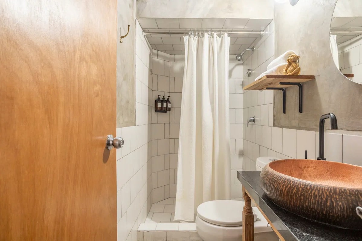 The bathroom features a modern design, with textured concrete walls and white tile accents. A circular mirror hangs above a wooden sink with an organic shape, and a shower is enclosed with a white curtain. Natural light may be visible from an unseen source.