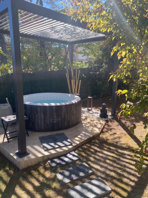 A private outdoor area features a hot tub under a shaded structure. Natural light filters through the slatted roof, creating interesting patterns on the ground. Stone pavers lead through the garden, alongside greenery and decorative elements, enhancing the tranquil setting.