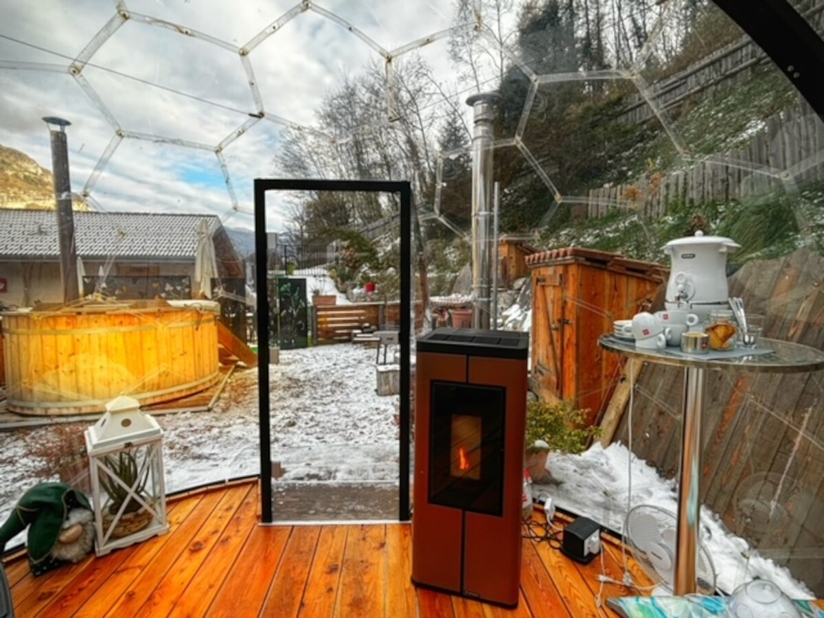 A transparent geodesic dome is seen, showcasing a wooden floor and views of a snowy outdoor area. A glass door leads to a wooden deck, with a round hot tub and seating in the background. A modern stove is positioned inside, complemented by a tea set on a table.