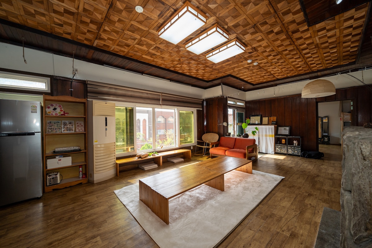 A spacious living area features a wooden ceiling with a geometric design, complemented by large windows allowing natural light. A low wooden table rests on a light rug, while a red couch adds a pop of color. A small kitchenette is visible to one side.