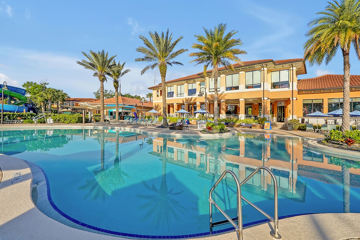 A large, inviting pool area is surrounded by lush palm trees and colorful landscaping. The pool's calm water reflects the sunny sky, while sun loungers and umbrellas provide shaded seating options. A spacious clubhouse is visible in the background, creating a relaxed resort atmosphere.