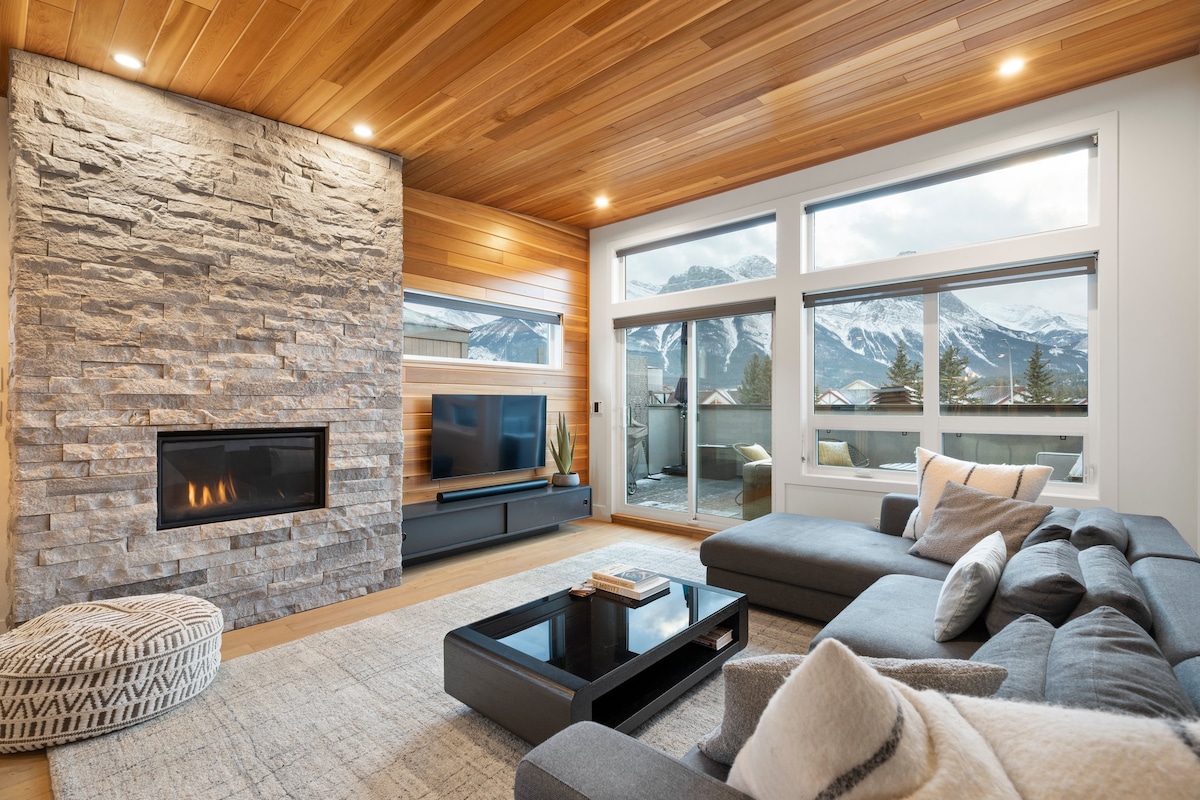 The spacious living area features a stone fireplace and large windows that frame mountain views. A comfortable grey sectional lounge sofa, a round ottoman, and a coffee table enhance the inviting atmosphere. Soft lighting highlights the wood-paneled ceiling and modern decor.
