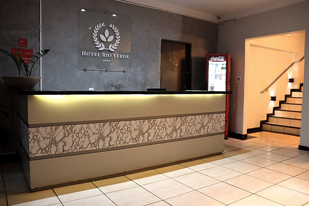 The reception area features a stone-patterned front desk with soft lighting and a logo on the wall. A vending machine is located to the side, and a staircase leads to the upper level. Neutral tiles create a welcoming atmosphere.