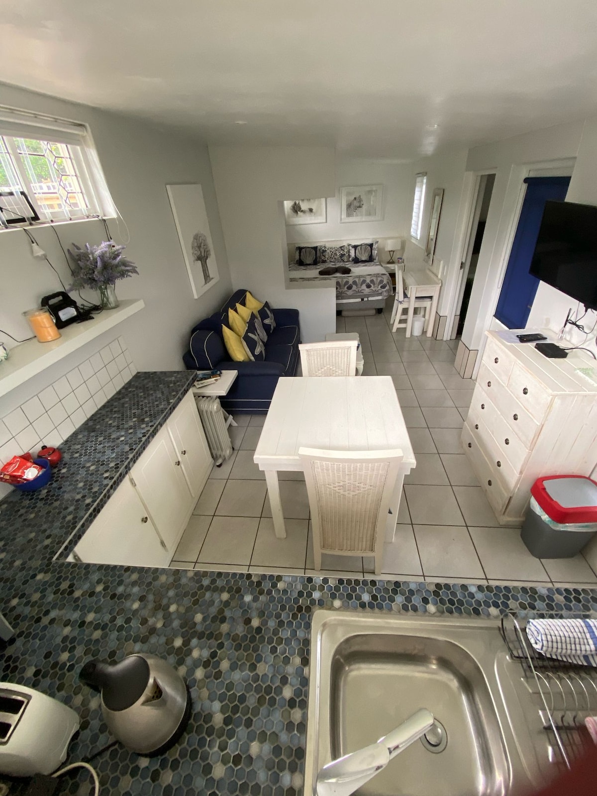 A well-equipped kitchen is seen with a tiled floor and sleek countertops. A dining table with matching chairs is positioned next to a cozy seating area. Light walls create a spacious feeling, while a television mounted on the wall offers entertainment options.