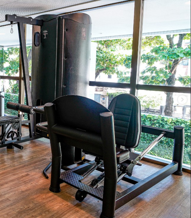 A well-equipped gym space is shown, featuring dual weight machines designed for strength training. Large windows provide natural light, offering views of greenery outside. The area is maintained with wooden flooring, creating a balanced and functional workout environment.