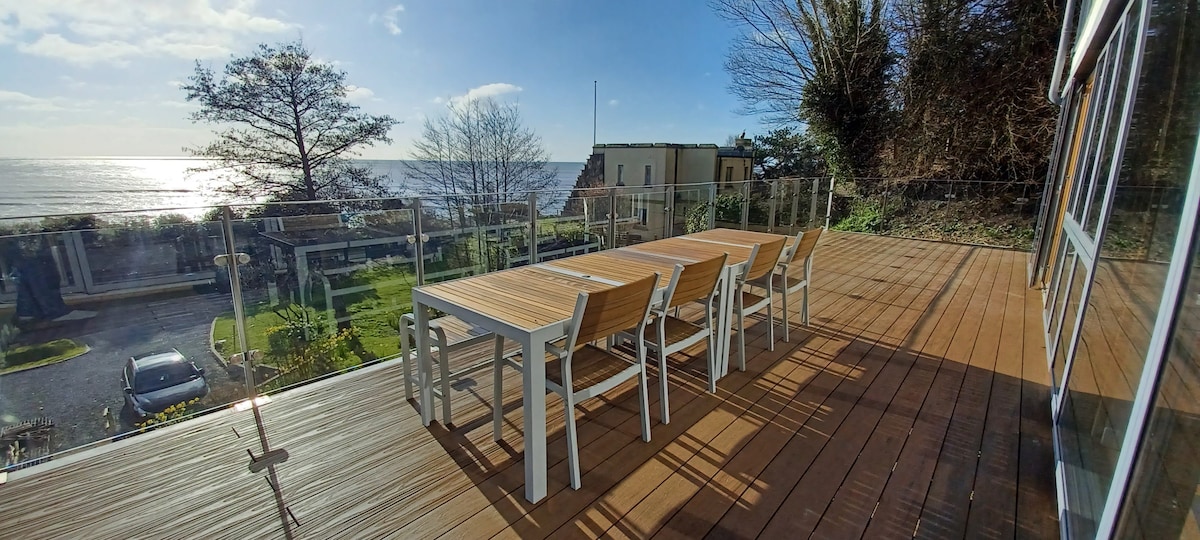 A spacious balcony features a wooden deck with a large table and six chairs, offering direct views of the sea and Rye Bay. Glass railings enhance the view, while greenery and the beach can be seen in the background.