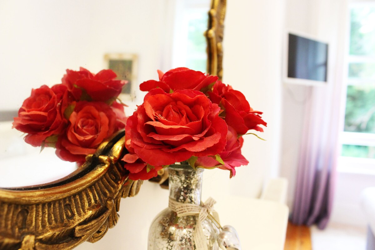 A glass vase filled with red roses is positioned against a vintage-style gold-framed mirror. The soft reflection of the flowers can be seen in the mirror, adding a touch of elegance to the room. A television is mounted on the wall in the background.