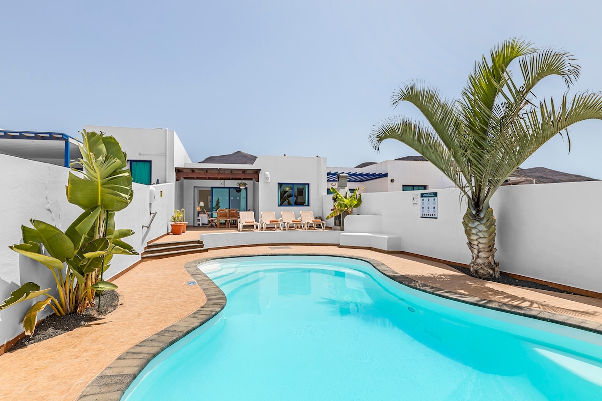 The image features a private swimming pool surrounded by terracotta tiles. Lush green plants, including a tall palm, add a tropical feel. Sun loungers are arranged near the pool, with a white-walled villa visible in the background against a clear blue sky.