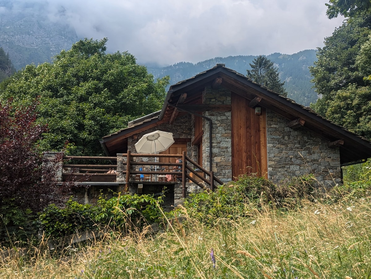 A charming stone house is set against a mountainous backdrop, surrounded by lush greenery. The wooden accents enhance its rustic appeal, and a private terrace is visible, complete with an umbrella. The natural landscape features wild grasses, contributing to the tranquil environment.