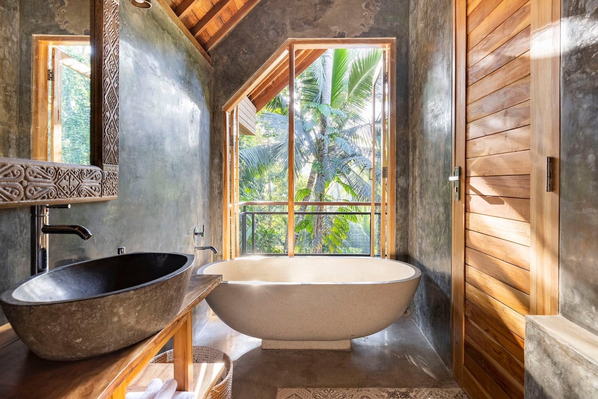 A modern bathroom features large windows that frame views of lush greenery. A freestanding tub rests beside the window, while a sleek, dark stone sink is positioned against a textured wall. Natural wood accents are present in the door and decor, enhancing the tranquil ambiance.