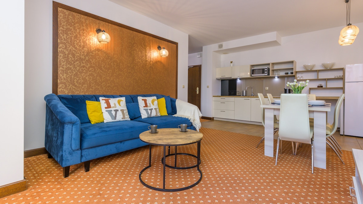 The open living area features a blue sofa adorned with decorative pillows, accompanied by a round coffee table. Nearby, a dining space with white chairs is visible. The kitchen includes modern appliances and neutral cabinetry, while a patterned rug adds texture to the floor.
