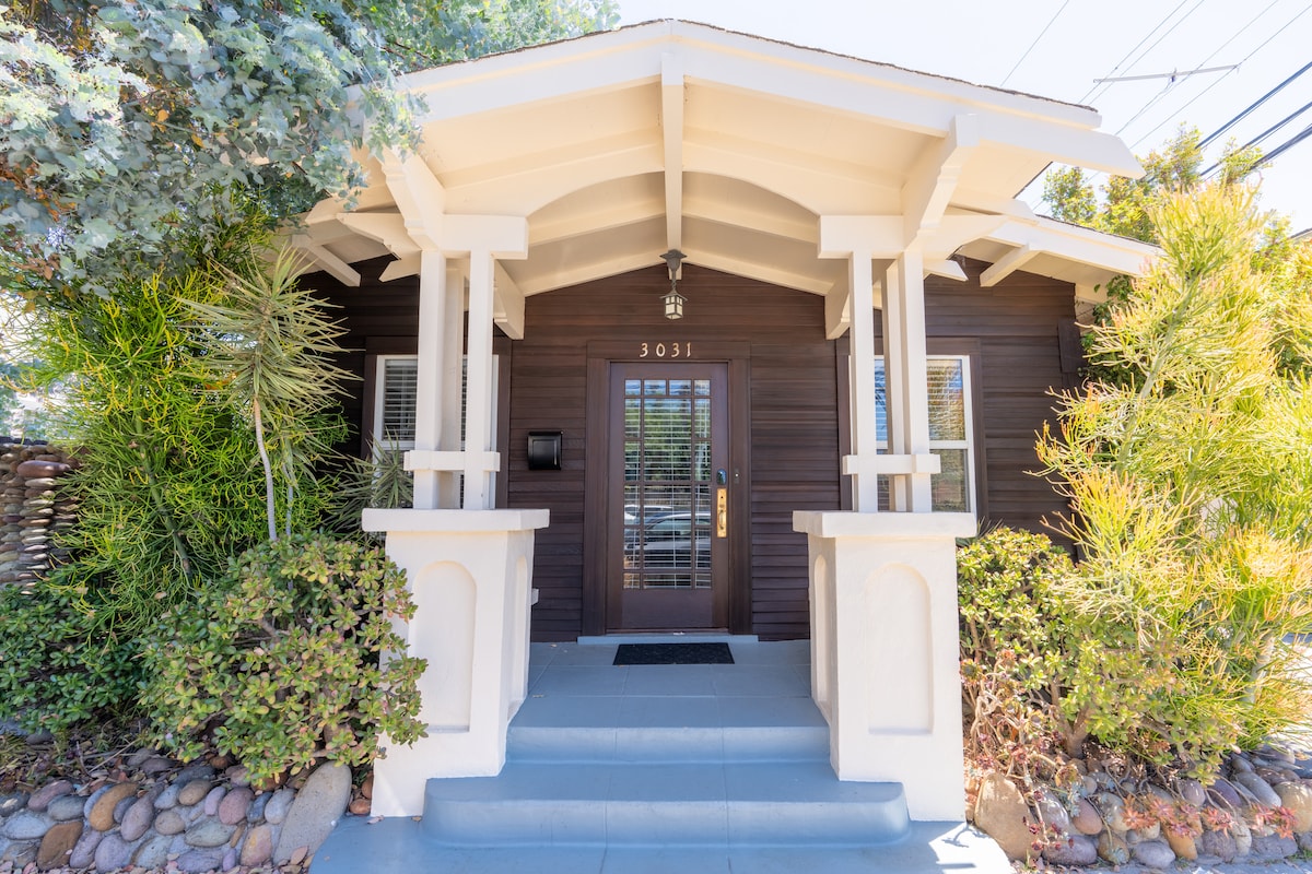 The entrance to the bungalow features a welcoming porch with two columns and a dark wooden front door. Lush greenery surrounds the entrance, including various shrubs and plants, while a smooth path leads to the door.