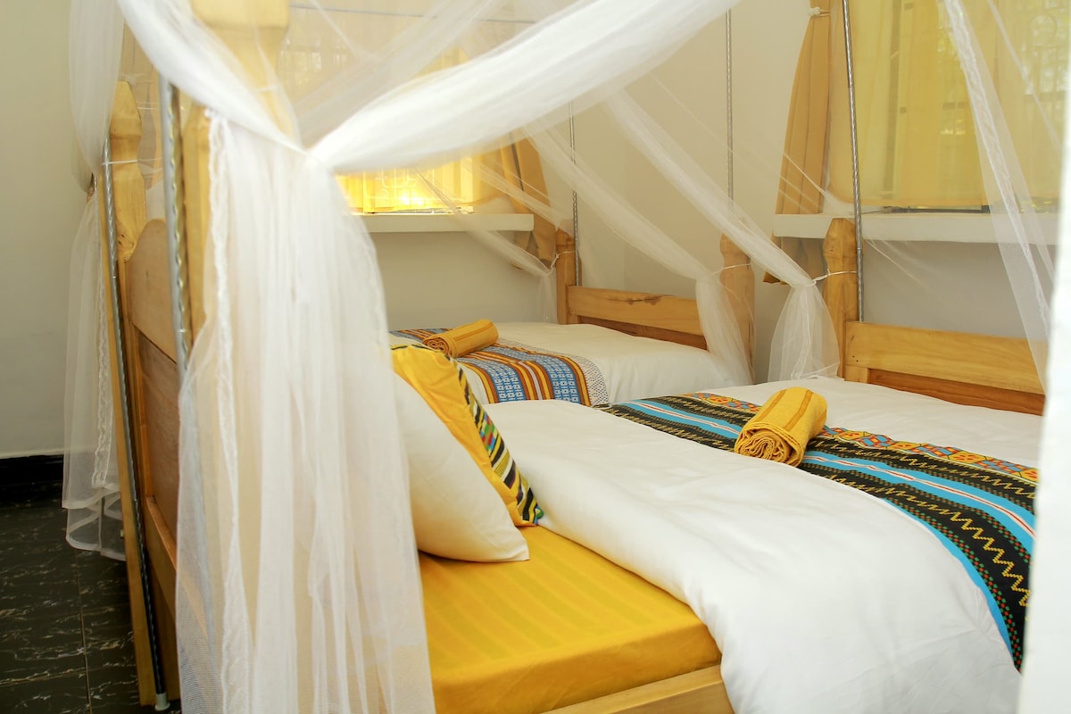 Two spacious beds are positioned underneath a delicate white mosquito net, creating a serene sleeping environment. Bright yellow accents on the bedding and decorative pillows add warmth, while natural light filters through nearby windows, softly illuminating the room.